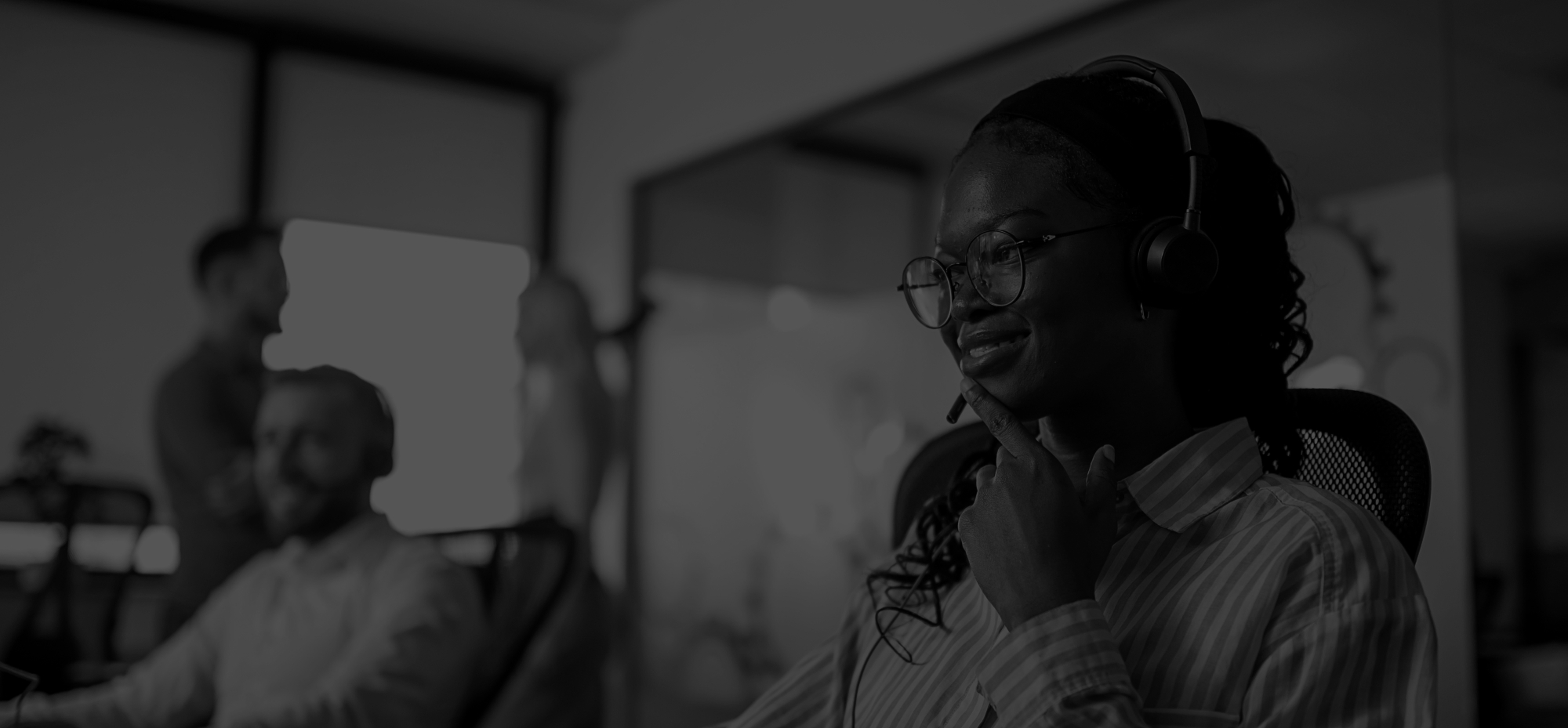 Smiling black female customer support agent wearing headset is working on computer in a modern office while colleagues are talking in the background